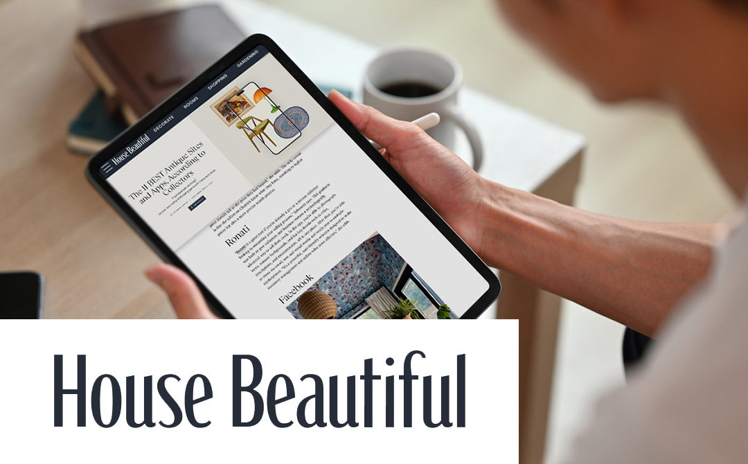 A person holding a tablet displaying the House Beautiful website's article about antique sites and apps. The tablet shows part of the article with visible headings including 'Ronati' and other furniture marketplace information. There's a coffee cup beside the tablet on a white surface with notebooks or journals nearby. At the bottom of the image, the House Beautiful logo appears in large dark text against a white background. The image illustrates someone researching online antique shopping resources from a March 2025 article about the best platforms for collectors.