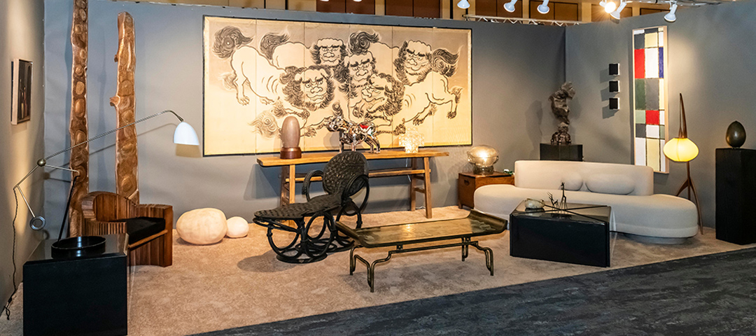 Interior display at JC Studios featuring a mid-century modern living room setup with a dramatic curved white sofa, glass coffee table, and sculptural black chair. The space showcases a large Japanese-inspired folding screen with lion-dog figures as the focal backdrop, flanked by wooden sculptural elements and colorful stained glass panels. The curated display combines contemporary furniture with artistic elements typical of modernist design exhibitions at Modernism Week.
