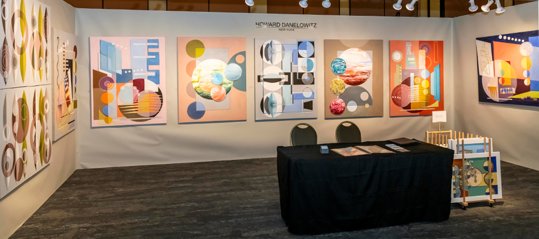Gallery exhibition space for Howard Danelowitz from New York, featuring a collection of colorful geometric abstract paintings arranged on white walls. The artworks display bold compositions with circles, rectangles, and organic shapes in vibrant colors including orange, blue, yellow, and pink. In the center of the space is a black-draped table with information materials and two chairs behind it. A small display rack with additional artwork stands to the right. The gallery name 'HOWARD DANELOWITZ NEW YORK' is visible above the central wall display.
