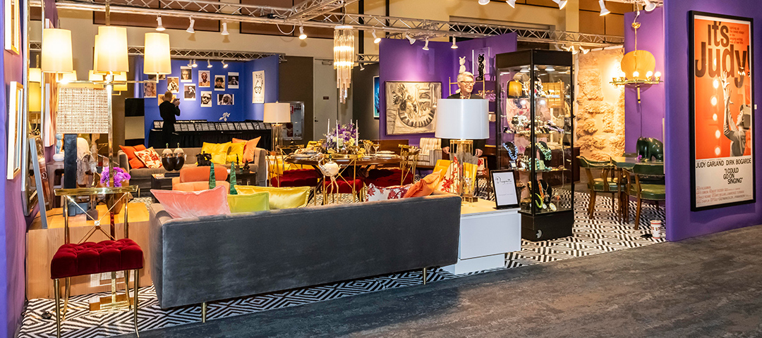 Vibrant showroom display of Dragonette Ltd at Modernism Week, featuring a colorful, eclectic mid-century modern interior with bold purple walls and geometric patterned flooring. The space showcases luxurious furniture including a gray velvet sofa, colorful accent pillows, and brass accents throughout. Various design objects, lighting fixtures, and decorative items are artfully arranged, with a striking orange 'It's Judy!' poster visible on the right wall. The exhibition combines Hollywood glamour with modernist design elements in a theatrical, gallery-like setting.