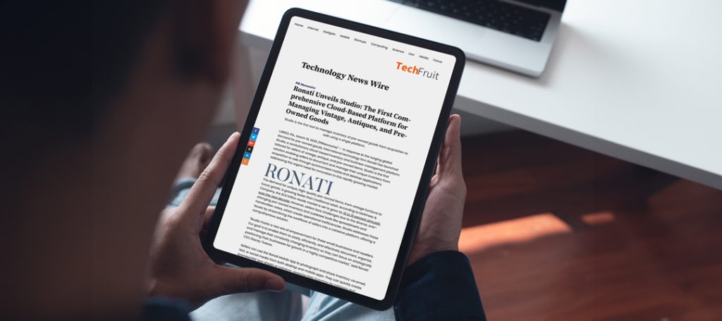 "A person holding a tablet displaying a TechFruit news article titled 'Ronati Unveils Studio: The First Comprehensive Cloud-Based Platform for Managing Vintage, Antiques and Pre-Owned Goods.' The tablet shows the TechFruit logo in orange at the top right, with the main article featuring a large 'RONATI' heading in the middle of the page. The tablet is being held by two hands against a wooden surface, with part of a laptop visible in the upper portion of the image. The article appears to be discussing Ronati's new platform for sellers of vintage and antique items.