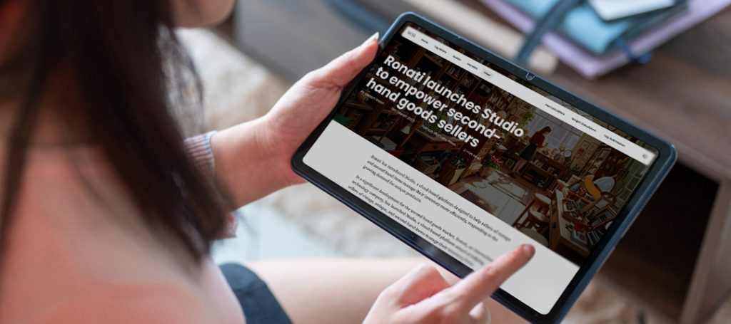 A person holding a tablet displaying a news article with the headline 'Ronati launches Studio to empower second-hand goods sellers.' The tablet shows what appears to be a vintage or antique shop in the background image of the article, with various furniture and decor items visible. The person is holding the tablet with both hands, with one finger touching the screen. Only the person's hands, part of their arm with a light-colored sleeve, and shoulder are visible in the frame. The setting appears to be indoors with a neutral background.