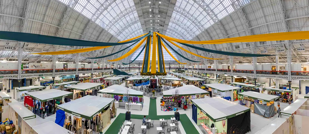 Panoramic view of Olympia's Grand Hall during a fair, showing the Victorian glass-roofed venue filled with white exhibition stands arranged in organized rows. Colorful fabric draping hangs from the central ceiling point, and visitors browse between the booths on the main floor.