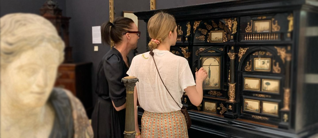 Two women examining an ornate black cabinet with multiple compartments containing decorative objects and artwork. A classical marble bust is visible in the foreground, representing the type of fine antiques and art pieces featured at the fair.