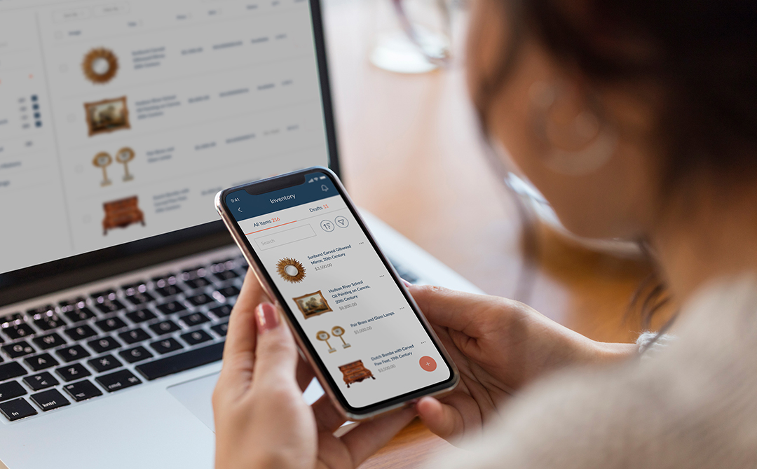 Antique dealer using Ronati Studio mobile app on smartphone to manage inventory, with laptop showing desktop version of the platform in background.