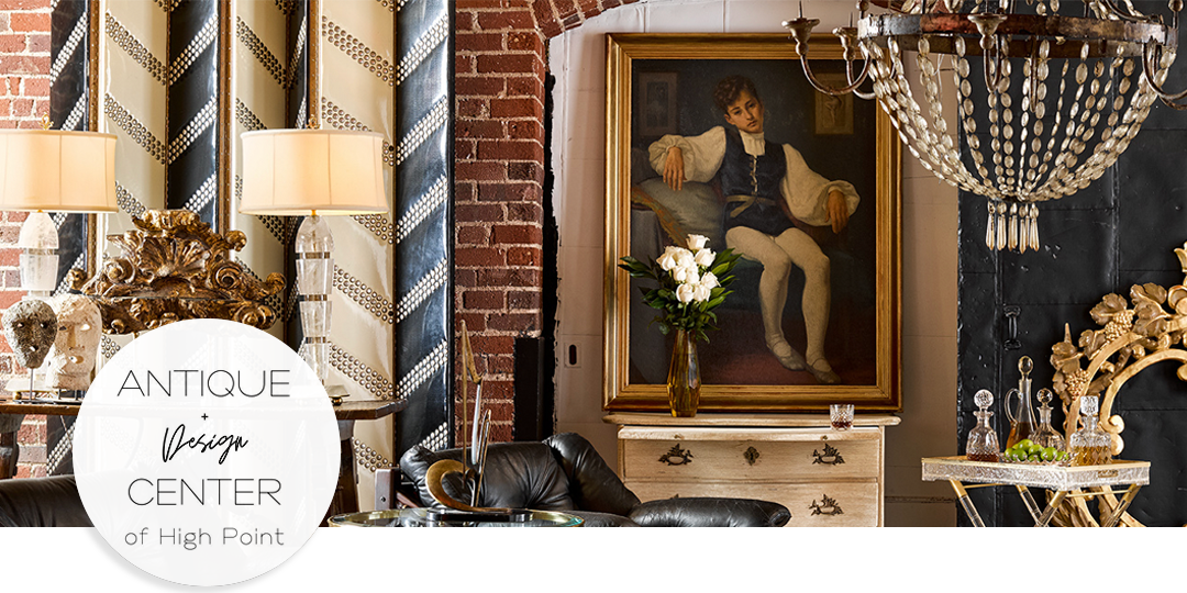 Eclectic interior at the Antique & Design Center of High Point featuring vintage furniture, gilded mirrors, and a portrait against an exposed brick wall.
