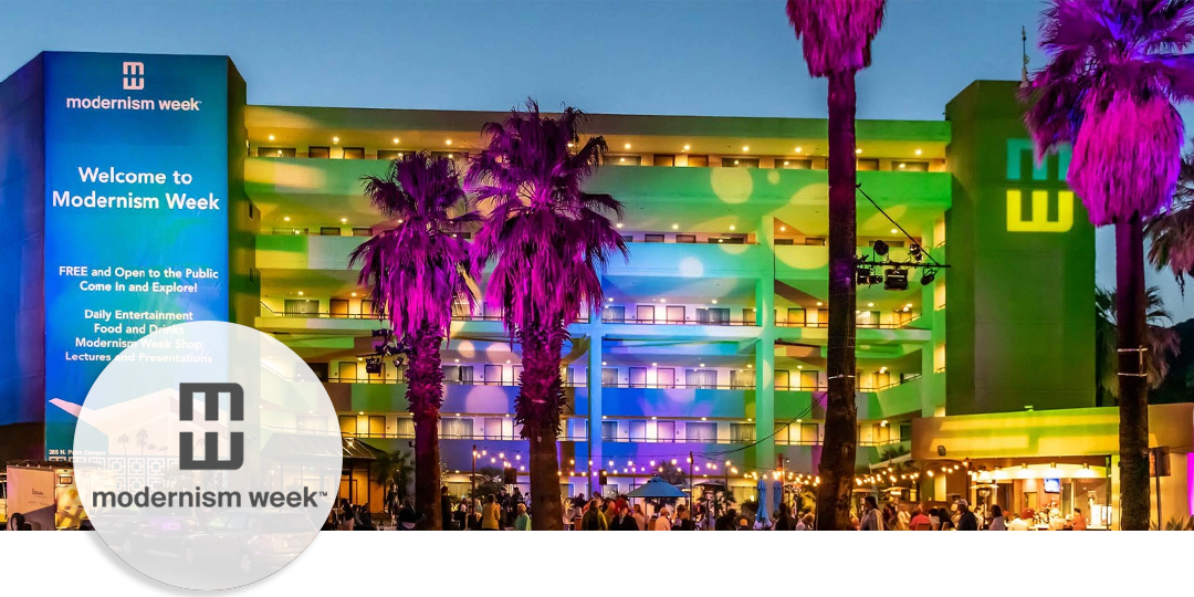 Night view of Palm Springs Modernism Week with mid-century building illuminated in colorful lights and visitors gathered beneath palm trees.