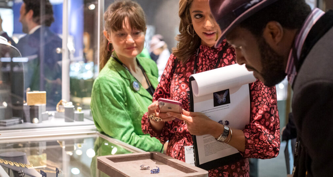Two women examining antique jewelry at a fair booth while speaking with the dealer — highlighting the personal connection of fairs.