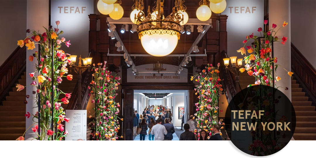 Floral-lined entry to TEFAF New York at the Park Avenue Armory, with crowds entering beneath a grand chandelier for the prestigious art and antiques fair.